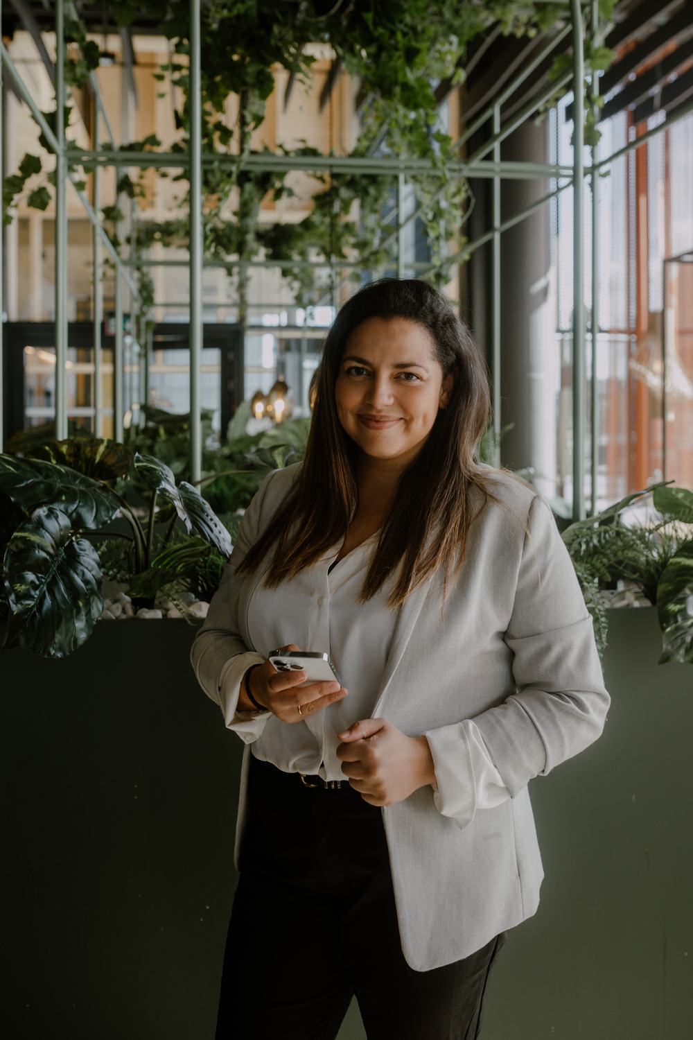 Ylonka Kaupang i lys blazer med mobilen i hånden, foran en grønn plantevegg